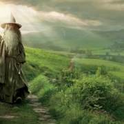 Gandalf the Grey from The Lord of the Rings stands on a stone path in the lush green hills of the Shire. Wearing his iconic pointed wizard hat and gray robes, he holds his wooden staff while overlooking the idyllic countryside. Sunlight breaks through clouds in the background, illuminating the pastoral landscape of rolling hills and distant fields that stretches toward the horizon. The scene captures Gandalf as the wise mentor figure at the beginning of the hero's journey.
