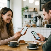 Two people at a coffee shop table, both smiling and holding phones showing completed payment transaction checkmarks, with coffee and pastries between them