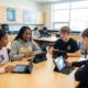 Four high school students collaborating around a table in a science classroom, each working on tablets with protective cases, with Biology, Chemistry, and Physics posters visible on the walls and additional students working at tables in the background